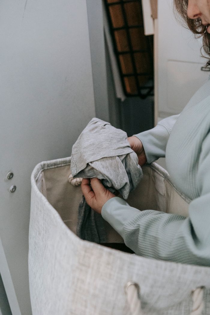 Crop anonymous female female choosing dirty clothes from wardrobe in bag while standing in light room during household routine routine at home
